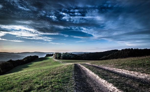 Germany, Westerwald. 
An unpaved field, forest and meadow path leads over the heights of the Westerwald. Some distance to the left and right we can see low mixed forest, with morning mist billowing in the valleys behind. The sky above with interesting cloud formations in all shades of blue and gray.  

Deutschland, Westerwald. 
Ein unbefestigter Feld-, Wald-, Wiesenweg führt über die Höhen des Westerwaldes. In einiger Entfernung sehen wir links und rechts niederen Mischwald, in den Tälern dahinter wabert Morgennebel. Der Himmel darüber mit interessanten Wolkenformationen in allen Blau- und Grautönen.