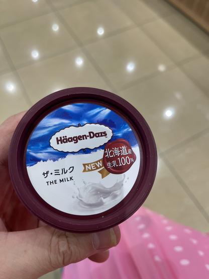 A person's hand holds a tub of Häagen-Dazs ice cream labeled "ザ・ミルク" (The Milk) with a "NEW" tag and a note indicating it is made from 100% Hokkaido milk. The background