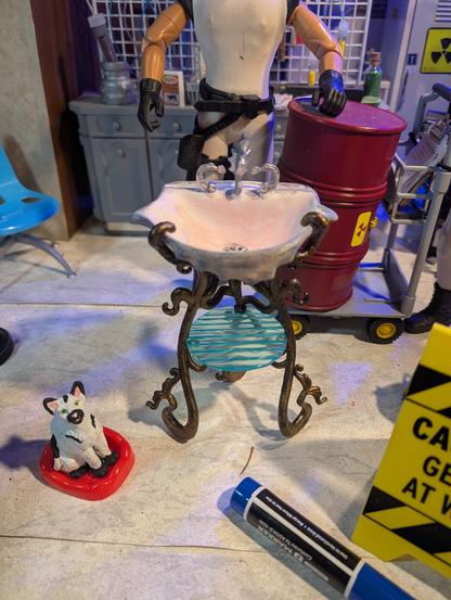 The tabletop set where I shoot most of my "Minions at Work" photo cartoons for Pulphouse Fiction Magazine. A black on white Minions cat sits in a cat bed at lower left. Also in the foreground are a felt marker (for scale) and just on the edge, a yellow floor tent-sign that reads "CAUTION: GENIUS AT WORK," thought you can't actually read it in the picture. Behind this Minion No. 1 (seen only from shoulders down) standing leaning on a barrel of radioactive sludge riding on a hand truck. A chemical laboratory bench is in the background.  In the middle is a steampunk sink, with an artfully curved gold-tone base that also supports a round, low, shelf apparently made of rippled blue glass. The sink bowl appears to be made from a giant clam shell. The faucet is shaped like a sea horse, and the curved handles are fish, heads down, tails arched over backs.