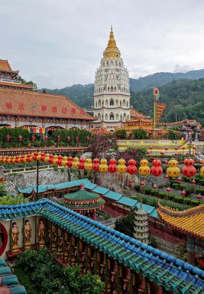 A high-angle, slightly aerial shot of a Buddhist temple complex. The central feature is a tall, white pagoda with a gold spire, rising in the middle ground. The complex includes multiple buildings with traditional East Asian architecture, featuring orange tiled roofs and intricate detailing. Rows of red and yellow lanterns line the walkways and gardens, adding vibrant color. Lush greenery surrounds the temple buildings, with a forested hillside visible in the background. The sky is overcast, suggesting a cloudy day.