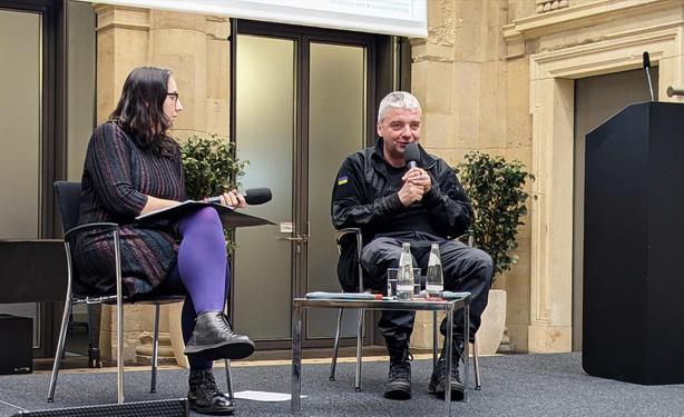 Olek­san­dra Bienert und Maxim Butkewitsch sithen rechts und links von einem niedrigen Tisch in Richtung Publikum. Beide halten Mikrofone, er spricht. Er hat sehr kurze hellgraue Haare und trägt einen dunklen Pullover mit einem kleinen blaugelben Aufnäher am Oberarm, dunkle Hosen und Stiefel. Im Hintergrund Wände aus beigem Sandstein.