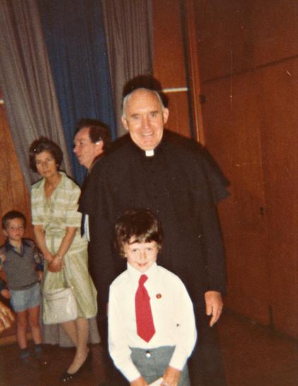A scan of a colour photo taken in the late 1970s. We're in what might be a school hall. The walls are panelled in warm-toned sheets of wood and the wall directly ahead of us has huge curtains tied back, the outer two in grey, the middle one in blue. A couple of adults and a child are standing in the background, near the curtains, but the focus of this shot is on a young boy and a priest. The boy is around seven or eight years old, wears a white shirt, red tie, and light grey trousers. He has a red button on his shirt breast pocket and is holding a sheet of paper in his hands barely in view at the bottom of the shot. He has unruly dark hair and a lopsided smile that almost conceals looks sinister. The priest wears all black with the small white collar, is mostly bald, and is smiling. Flash from the camera gives him a dark shadow behind. The photo is at a slight tilt adding to the mild sense of menace.