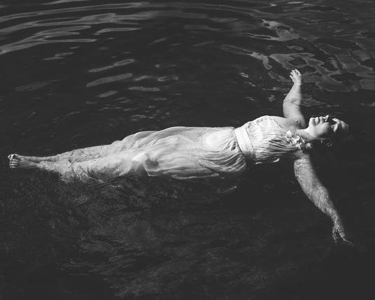 The image shows a person floating on their back in water, wearing a long, flowing dress. The photo is in black and white, and the person's arms are outstretched.