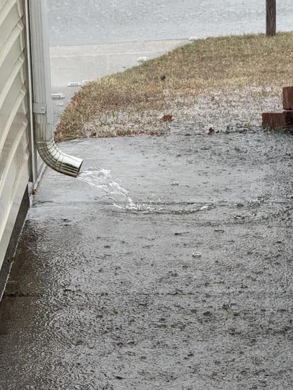 The image shows water flowing from a downspout onto a wet surface, likely due to rain. The water is pooling and running along the ground. There's a patch of grass and a sidewalk or path visible next to the downspout. It appears to be a rainy or wet day.
