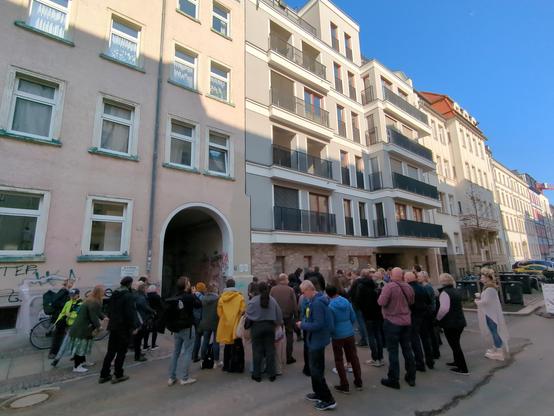 Circa 50 Menschen stehen vor 5-stöckigen Mehrfamilienhäusern auf der Straße, links ein Altbau mit Toreinfahrt, rechts ein moderner Neubau
