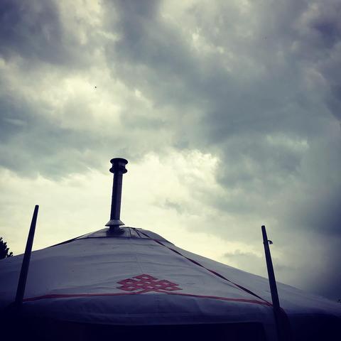 Das Bild zeigt eine Jurte gegen einen bewölkten Himmel. Die Jurte hat ein weißes Dach mit einem roten, ornamentalen Muster darauf. In der Mitte des Daches befindet sich ein Schornstein. Zwei lange Stäbe ragen an den Seiten des Daches in den Himmel. Der Himmel ist grau und voller Wolken.