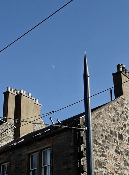 A colour photograph of a stone building in Leith. At both the left and right hand edges are chimney stacks. Below the right chimney is a stone wall and the gable end, strongly illuminated by the sun. The front of the building, from the centre to the left, is in deep shadow.

There is a pointed pole in front of the building towards the left supporting cables providing power to the trams, and two such cables slant diagonally from top right to bottom left of the picture. 

Behind the building and filling much of the picture is the cloudless, intense blue sky, in the middle of which the half-moon is just visible.