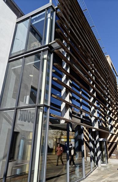 A colour photograph of a modern office building in Leith. The building occupies most of the picture. The building is glass-fronted, with vertical steel supports, and horizontal steel struts which do not appear to have a function. 

The strong sunlight hitting the building causes alternating bright and shadowed areas, the shadows caused by the struts. 

The reflections of people walking on the street can be seen in the glass, although the people themselves are not in the photo. 

Above the building to the right is a patch of intense blue sky.