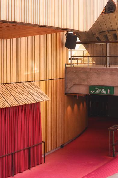 The image shows an interior space with a modern design. There are wooden panel walls and a red carpeted floor. A red curtain is visible on the left, and a sign indicating "Toilets and Bar" is on the right. The space has a clean, architectural look with concrete and wood elements.