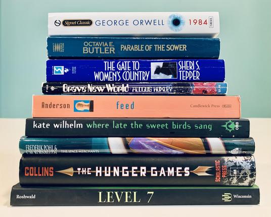 Pictured is a stack of books including George Orwell's 1984, Octavia Butler's Parable of the Sower, Sheri Tepper's The Gate to Women's Country, Aldous Huxley's Brave New World, M.T. Anderson's Feed, Kate Wilhelm's Where Late the Sweet Birds Sang, Frederik Pohl and C.M. Kornbluth's The Space Merchants, Suzanne Collins's The Hunger Games, and Mordecai Roshwald's Level 7.