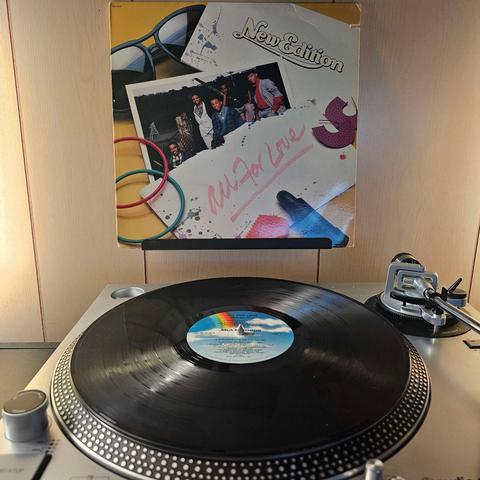 A vinyl record sits on a turntable. Behind the turntable, a vinyl album outer sleeve is displayed. The front cover shows various items like sunglasses, bracelets, marker, photo, and ripped paper. In the photo are the members of New Edition looking at the camera.