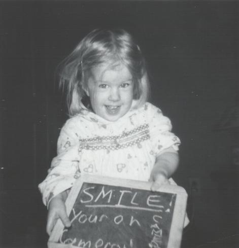 A black-and-white photo from the late 1960s of a little toddler girl in front of a dark background. Her hair is blonde and disheveled, and she is wearing pajamas. She is smiling adorably, while holding.a small chalkboard in front of her which reads, "SMILE! ...You're on Candid Camera."
