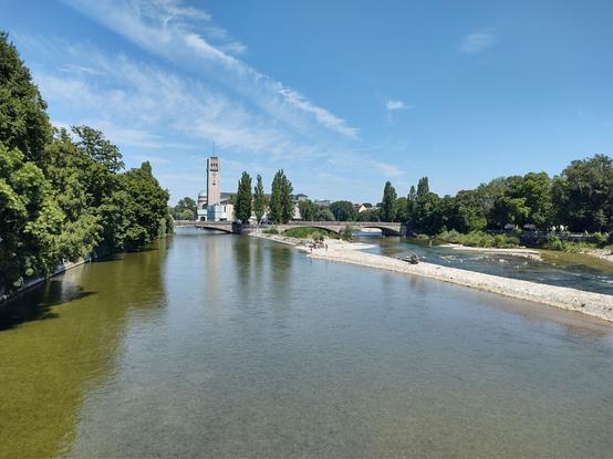 ChatGPT:

Das Bild zeigt eine Flusslandschaft an einem sonnigen Tag. Ein ruhiger Fluss mit klarem Wasser erstreckt sich durch die Szenerie, umgeben von üppigem, grünem Baumbestand auf beiden Seiten. In der Mitte des Bildes führt eine sandige, helle Kiesbank in den Fluss hinein, auf der einige Personen spazieren oder sich aufhalten.

Im Hintergrund steht ein markanter Turm mit einer Uhr und einer kleinen Kuppel, der zu einem größeren Gebäudeensemble gehört. Eine Brücke überspannt den Fluss, verbindet die Uferseiten und führt in Richtung des Gebäudes. Der Himmel ist strahlend blau mit wenigen, leicht verstreuten weißen Wolken.