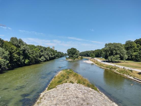 Das Bild zeigt eine idyllische Flusslandschaft an einem sonnigen Tag. Der Fluss teilt sich in zwei Arme, die eine kleine, mit Bäumen bewachsene Insel umschließen. Das Wasser ist klar und flach, und einige Menschen genießen es, im Fluss zu schwimmen oder auf den Kiesbänken zu entspannen.

Auf der rechten Seite verläuft ein Uferweg, auf dem Spaziergänger und Radfahrer unterwegs sind. Große, schattenspendende Bäume säumen das Ufer, während eine weitläufige Grünfläche im Hintergrund zu sehen ist. Auf der linken Seite ist dichtes, sattgrünes Gebüsch zu erkennen, das den natürlichen Charakter der Landschaft unterstreicht.

In der Ferne erhebt sich ein markanter Kirchturm über den Baumwipfeln, der ein architektonisches Highlight in der natürlichen Szenerie bildet. Der Himmel ist strahlend blau mit wenigen zarten Wolkenstreifen, was die sommerliche Atmosphäre unterstreicht.
