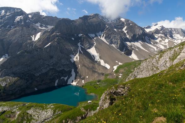 Blick von einem Berggipfel auf einen grünen Bergsee. Im Hintergrund ein paar Gipfel der Schweizer Alpen.