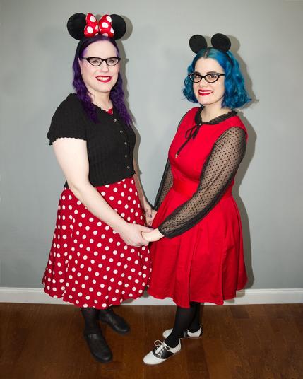 Two women stand side by side, wearing matching Mickey Mouse ears and vibrant, polka dot dresses, holding hands in a warm embrace.