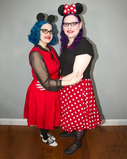 Two women, one with blue hair and another with purple hair pose together, wearing
Mickey and Minnie Mouse ear headbands and colorful vintage-style dresses.