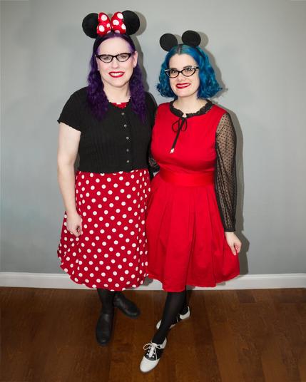 Two women, one with blue hair and another with purple hair pose together, wearing
Mickey and Minnie Mouse ear headbands and colorful vintage-style dresses.
