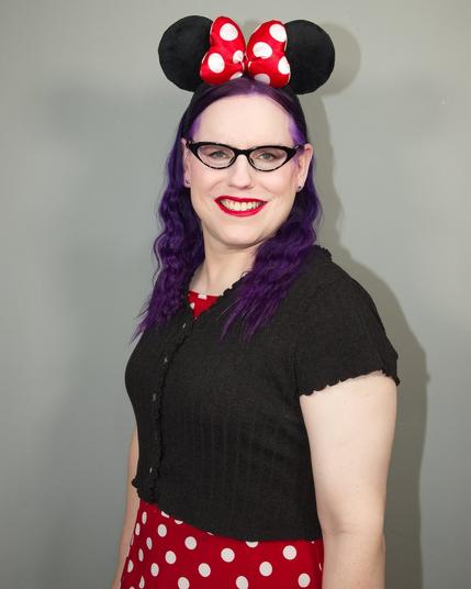 A woman with purple hair wears a black cardigan and a red polka dot dress, topped with black Minnie mouse ears and a red bow.
