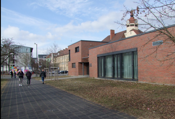 Plattenweg in ca. 5 m Breite mit einigen Fußgängern, beidseits Rasenflächen - drei Fußgänger nebeneinander nehmen etwas 2/3 des Weges in Anspruch