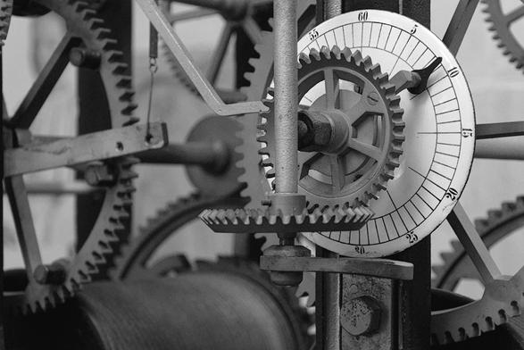 The image shows a close-up of an intricate clock mechanism, featuring interlocking gears and a dial with numbers. It highlights the detailed workings within the clock, including various size gears and mechanical linkages necessary for timekeeping.