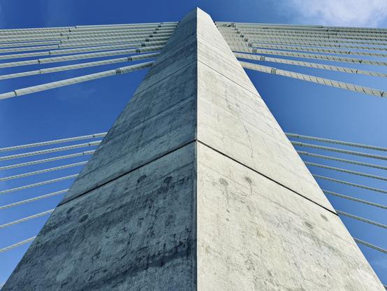 One side of a concrete suspension bridge tower rises up into the blue sky, as numerous white suspension cables extend outward and downward from it.