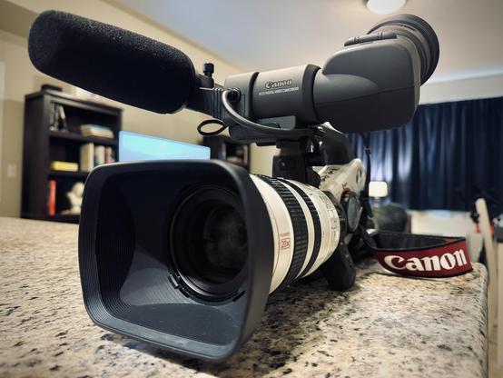 A large, white and matte black prosumer camcorder sits atop the counter of a residence, the red and black camera strap reading "Canon," and the front of the viewfinder reading, "Canon | 3CCD DIGITAL VIDEO CAMCORDER."
