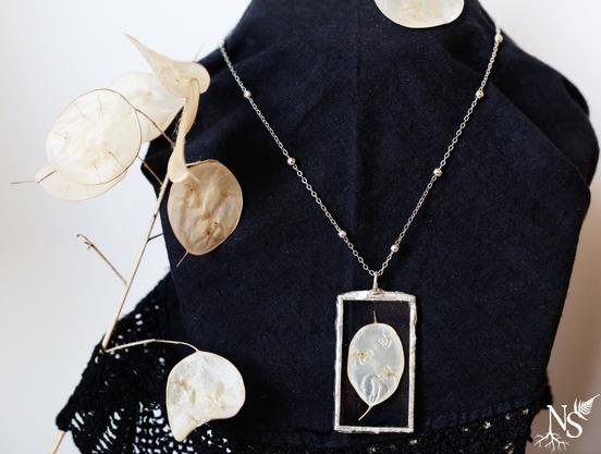 English A necklace stand covered with a black doily on which rests a silver necklace. It consists of a fine chain with pearls scattered along its length, and a vertical rectangular glass pendant containing a pearly white leaflet of plant origin. A branch with the same kind of leaflets rests carelessly on the side of the display to show where the plant part comes from.
•
Français Un présentoir à collier couvert d'un napperon noir sur lequel repose une collier argenté. Il est constitué d'une chaîne fine avec des perles disséminées sur sa longueur, et d'un pendentif rectangulaire vertical en verre contenant un feuillet blanc nacré d'origine végétale. Une branche avec le même genre de feuillets repose négligemment sur le côté du présentoir pour montrer d'où vient la partie de la plante.