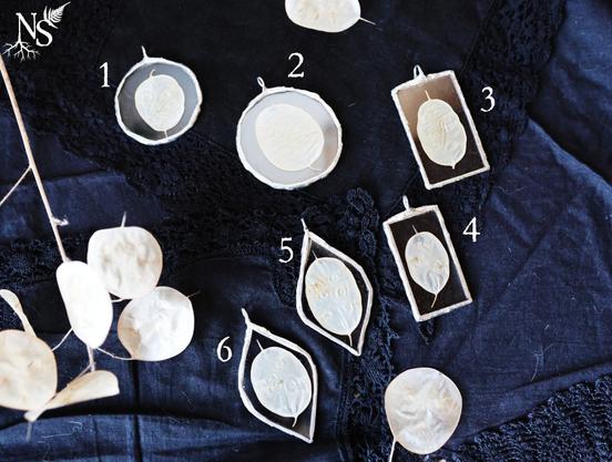 English Glass pendants containing pearly white lealets of plant origin rest on a background of black lace fabrics. The pendants come in a variety of shapes (round, vertical rectangular, vertical leaf). There are numbers next to each pendant so you can choose the one you prefer. A branch with the same kind of leaflets rests carelessly on its side to show where the plant part comes from.
•
Français Sur un fond de tissus noirs à dentelle reposent des pendentifs en verre contenant des feuillets blancs nacrés d'origine végétale. Les pendentifs ont des formes variées (rond, rectangulaire vertical, feuille verticale). Il y a des numéros à côté de chaque pendentif pour choisir celui qu'on préfère. Une branche avec le même genre de feuillets repose négligemment sur le côté pour montrer d'où vient la partie de la plante.