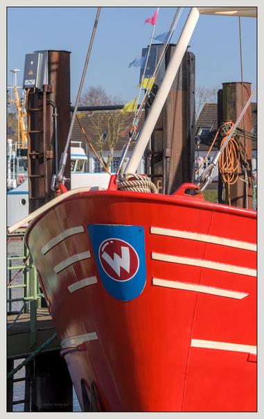 Nahaufnahme des Bugs eines roten Fischkutters mit einem blauen Emblem mit dem weißen Buchstaben „W“. Das Boot liegt am Kai, im Hintergrund sind Flaggen und verschiedene Gebäude zu sehen. Helles Sonnenlicht unterstreicht die leuchtenden Farben.

Close-up of the bow of a red fishing boat with a blue emblem with the white letter “W”. The boat is moored on the quay, flags and various buildings can be seen in the background. Bright sunlight underlines the vibrant colors.