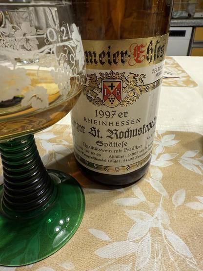 A bottle of 1997 Rheinhessen wine labeled "Kastanienhof St. Rochuskapelle Spätlese" is displayed next to a green glass with an etched grapevine design, set on a floral-patterned tablecloth