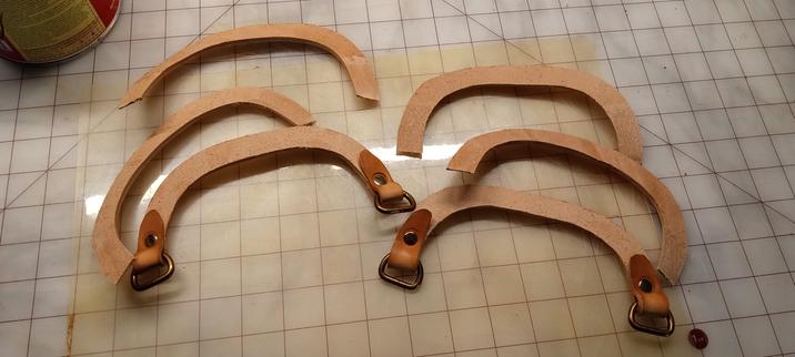 Six pieces of vegetable tanned leather, grain side up. They are shaped like horseshoes with the tips cut off. Two have longer ends that have been folded back onto themselves to hold brass "D" rings. These are riveted into place with the round end of the "D" inside the fold.