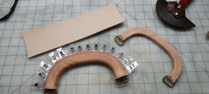 Two handles with a thin rectangular piece of vegetable tanned leather beside them. One handle has had leather wrapped around it and this leather is held in place with small, clear spring clamps. The other handle does not have a wrapping but shows signs of having been trimmed to round its shape. It should be understood by the composition of the photo that the rectangular piece of leather will be wound around the handle that  has not yet been wrapped.