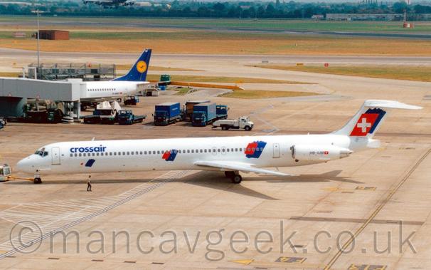 Side view of a white, twin engined jet airliner with the engines mounted on the sides of the rear fuselage, facing to the left but being pushed back to the right from it's gate by an almost-out-of-frame white truck attached to the nosewheel by a long yellow pole.
There are blue "CrossAir" titles on the upper forward fuselage, with red and blue splotches on the sides of the fuselage just afore and aft of the wing, repeated on the tail overlaid with a white cross.
The registration "HB-IUM" is on the side of the engine pods in black.
Concrete apron fills the foreground, with taxiways and a runway separated by grass in the background.
A grey airbridge stretches in to frame from the left, attached to a white, twin engined jet airliner with a blue and yellow tail.