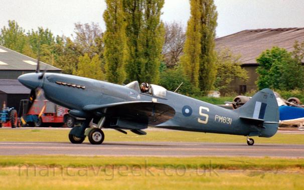 Side view of a World War 2-era single propellor-engined fighter plane taxiing from right to left on a tarmac runway.
The plane is a grey-blue, with white serial "PM631" on the rear fuselage, with a large white letter "S" slightly further forward.
There is a dark and light blue roundel just behind the cockpit, and a dark and light blue fin flash on the tail.
Green grass fills the foreground, with more grass leading up to a pair of hangars in the background, the one on the right behind trees.
Grey sky completes the picture