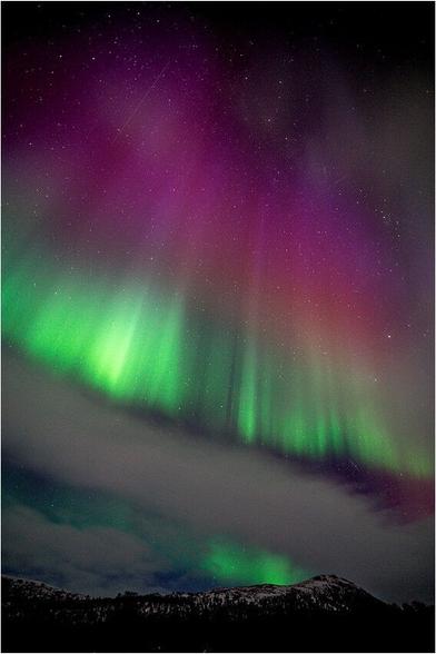 Polarlicht mit Verlauf von Rot (oben) nach Grün (unten) mit einem Wolkenband im unteren Bereich über schneebedeckten Bergen