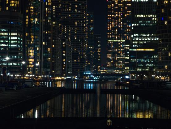 View of many skyscrapers around a central area of water at night, their lights reflecting down onto it. Near the front of the shot is a pair of locks holding the water in.