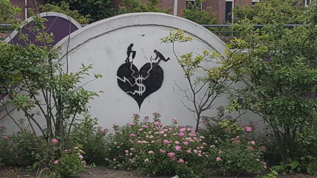Pink flowered plants and shrubs in front of a white, half circle concrete wall with black graffiti of a heart with a dollar sign on it being broken into pieces by a person with a jackhammer and one with a pickaxe.