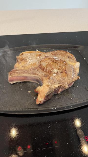 Una chuleta de ternera con hueso se está cocinando en una plancha caliente sobre una placa de inducción. La carne presenta un dorado en la superficie, mientras que el interior cerca del hueso aún muestra un tono rosado. Se pueden ver escamas de sal gruesa sobre la carne y pequeñas gotas de grasa en la plancha. Hay un ligero humo saliendo de la cocción, lo que indica que la carne se está sellando a alta temperatura. La encimera y la placa de inducción reflejan la luz del ambiente, mostrando un aspecto limpio y moderno.