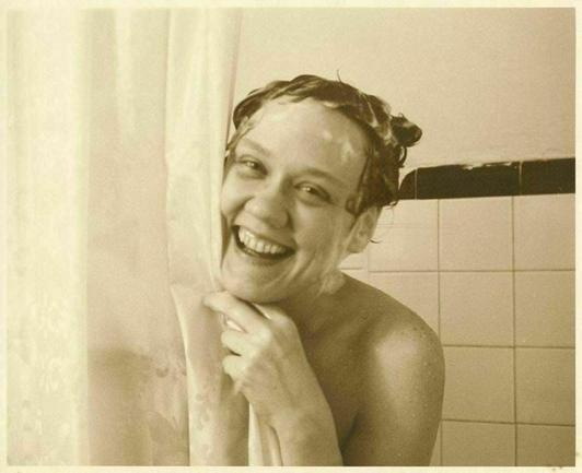 Woman poking her soapy joyful, smiling face from behind the shower curtain, along with one hand and one bare shoulder. She is being photographed by her lifelong partner.