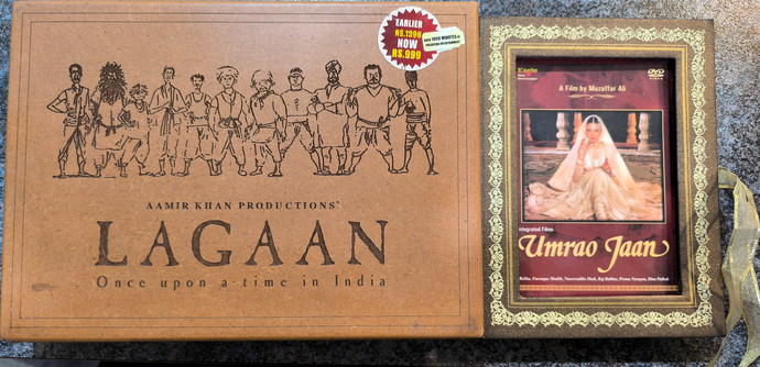 On the left, a wooden case with an engraved image of the core cast of the Hindi movie Lagaan, and the movie title "Lagaan Oce Upon a Time in India"  all engraved in the lid. Next to it, an ornamented DVD case for the movie Umrao Jaan. The centre image is the title character and around the edge of the case is a border that looks like emroidered fabric, with a lacy ribbon attached to the side of the case.