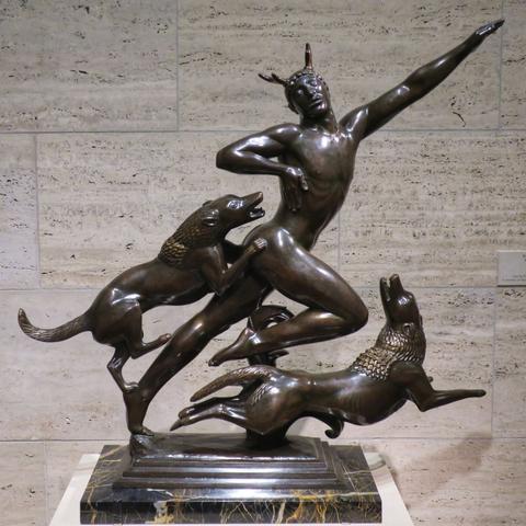 A sculpture in bronze of a man/ancient-god in a balletic pose with one leg raised, left arm outstretched as he races forward, pursued by two wolves bent on destroying him.