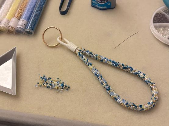 A bead-wrapped wristlet keychain on a bead board with a pile of bead soup in blues, gold,  yellow, and white. You can also see a bead scoop, a needle, and a bottle of superglue on the board.
