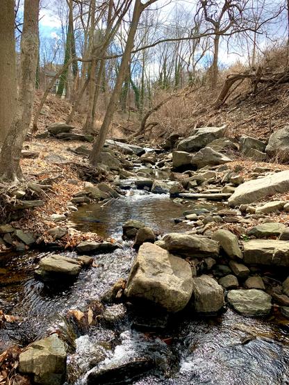 A small stream flows down the narrow valley, winding through a rocky landscape surrounded by woods. There are many rocks, both big and small, scattered in and along the stream.