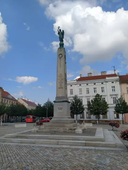 The image depicts a monument or statue situated in a square or public area. It features a tall column topped with a figure, possibly representing a woman holding something above her head, which appears to be an emblem or symbolic object. The inscription on the monument includes text that may commemorate a historical event or figure. In the background, there are buildings, trees, and possibly some vehicles, creating an urban atmosphere. The sky is partially cloudy.

Image Credits: Wikimedia / Chondrocladia / CC0