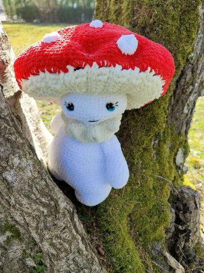 A crocheted plush toy resembling a mushroom with a red cap and white spots is nestled between the trunks of a tree. The toy has a white body, blue eyes with long eyelashes, and a small smile. The tree trunk is covered in moss, and the background shows a grassy area with some trees and sunlight filtering through.