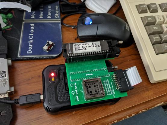 T48 EPROM programmer with an adapter board for 27C400/800/160/322 EPROM's that has a ribbon cable hooking into the side of it that jumps between the adapter and base board. There's a 27C160 EPROM chip in the ZIF socket with a marking on pin 2 (which will become pin 1 when inserted into the Amiga 500; actual pins 1 and 2 will be grounded). There's a piece of foil underneath a strip of tape covering the ultraviolet erasure quartz window on the chip. Written on the tape is "Kickstart 3.1 / Eat shit and die, Cloanto"