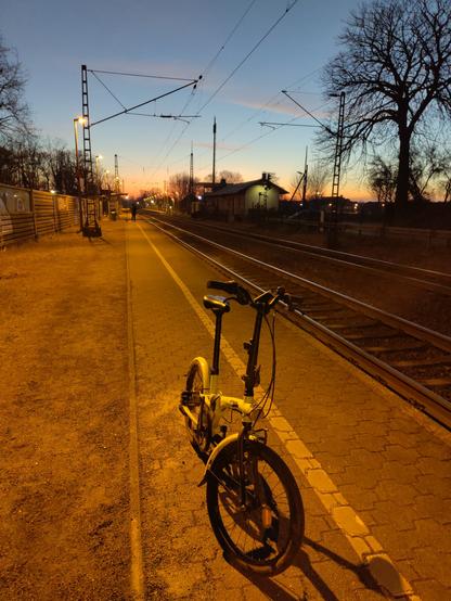 Bild eines Bahnhofs mit zwei Gleisen. Auf der gegenüber liegenden Seite ist ein altes Bahnhofsgebäude zu sehen. Am unteren Rand in der Mitte steht ein Klapprad. Der Bahnsteig ist in orangenes Licht getaucht. Im Hintergrund blauer Himmel mit aufgehender Sonne am Horizont.
