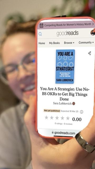 An iPhone portrait selfie, with me blurry in the background, holding my phone up in front of the camera in the foreground (in focus) showing a screen cap of the You Are A Strategist book page on GoodReads.com!
