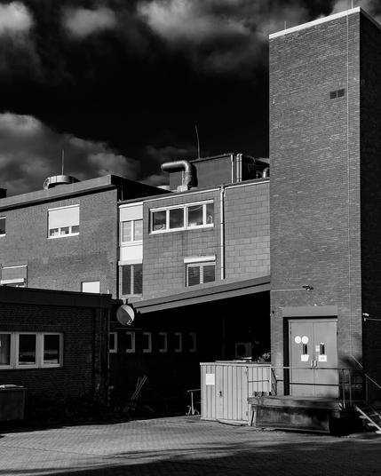 In dramatischem Schwarzweiß präsentiert sich ein Industriekomplex mit markanten Lüftungsrohren und Fenstern, während Wolken am Himmel für eine düstere Stimmung sorgen. Die klaren Linien und Backsteinfassaden verleihen dem Gebäude einen rauen, urbanen Charme.